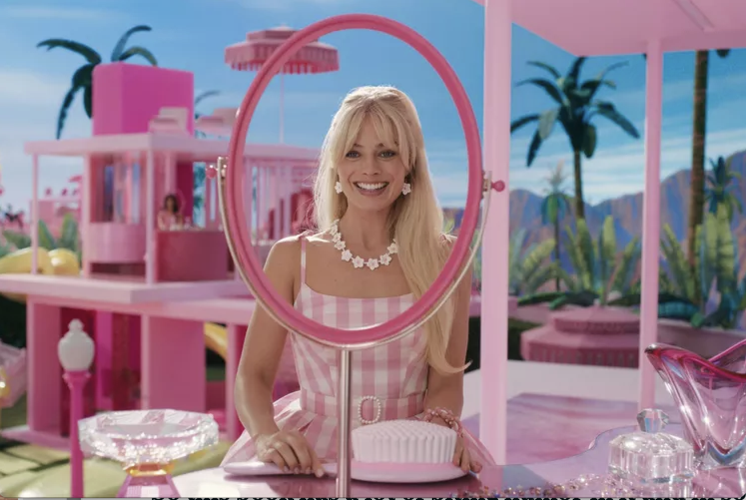 Margot Robbie as Barbie sits at a pink vanity, smiling into a big round mirror.